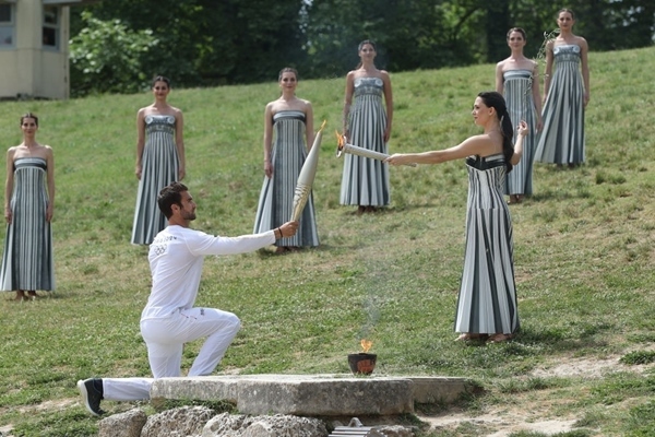 扮演最高女祭司的演員瑪麗·米娜（前右）點燃第一棒火炬手——希臘運動員、東京奧運會男子賽艇項目金牌得主斯特凡諾斯·恩圖斯科斯手中的火炬。新華社記者 趙丁喆 攝