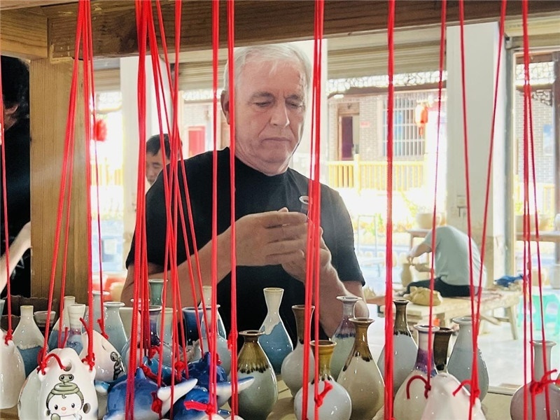 鄉(xiāng)愁如同一根根紅線，將游子與故鄉(xiāng)永遠(yuǎn)地牽連到一起?；逄账囄幕瘎?chuàng)意一條街的母氏陶藝館內(nèi)，紅線吊著陶瓶，思緒游走萬里。摩爾多瓦“信息標(biāo)簽”通訊社社長亞歷山大·塔納斯端詳著館內(nèi)展示的陶藝作品，并挑選出心儀的小擺件帶回摩爾多瓦，也帶走花茂村的“鄉(xiāng)愁”。（張亞欣攝）